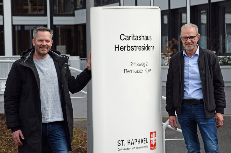 Zwei Männer stehen. In der Mitte ein großes Schild mit der Aufschrift: Caritashaus Herbstresidenz.