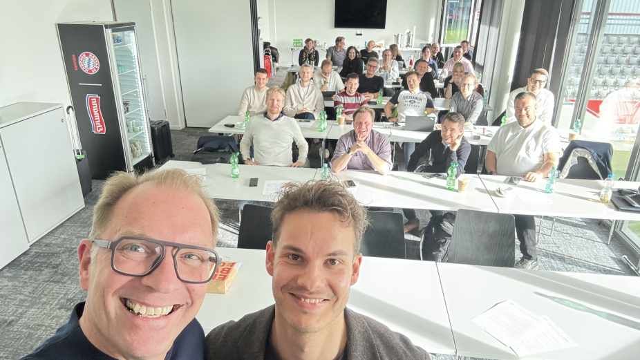 André Weinert und ein Mann lächeln in die Kamera. Im Hintergrund ein Seminarraum mit Menschen.