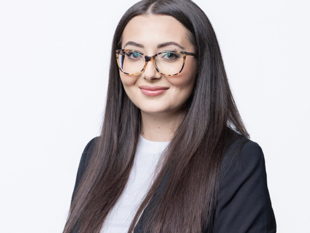 Mia Angelov trägt ihr langes, glattes, dunkelbraunes Haare offen und eine große braune Brille. Sie lächelt leicht in die Kamera. 