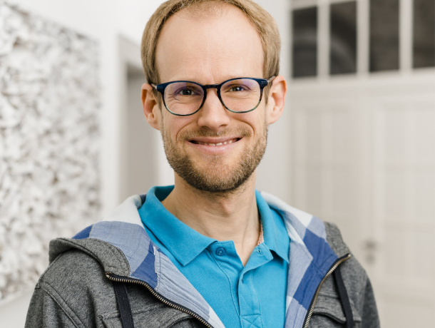 Daniel strahlt mit einem breiten Lächeln in die Kamera. Er trägt kurzes, mittelblondes Haar, eine dunkle Brille und einen Dreitagebart. 