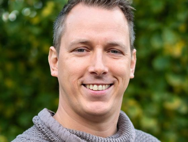 Marcel lächhelt mit kurzem Haar in einem grauen Pullover mit Rollkragen vor einem grünen, natürlichen Hintergrund.